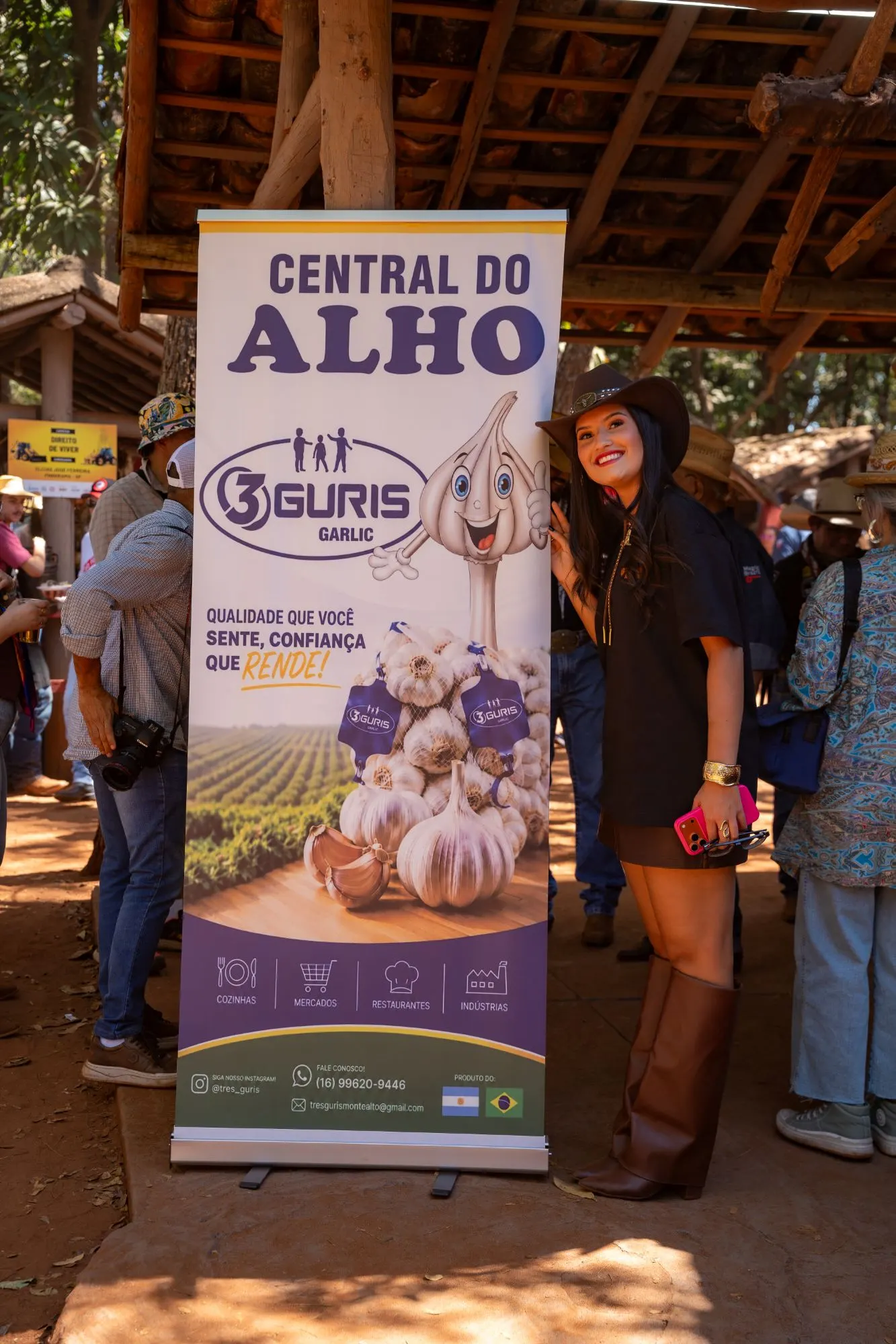 Cobertura de evento da Três Guris: convidada de chapéu sorrindo ao lado do banner “Central do Alho”, com fotógrafo e público ao redor